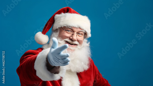 Old man in Santa Claus costume smiling and presenting or pointing at something isolated on blue background, Christmas background with copy space.
