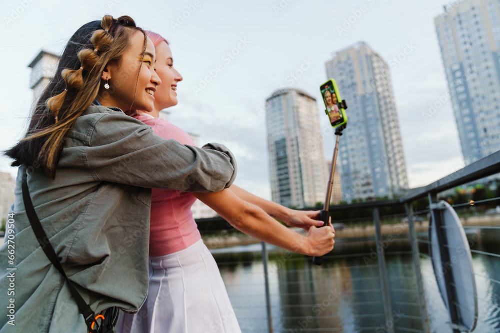 Two girls making video with smartphone and selfie stick while standing on city waterfront