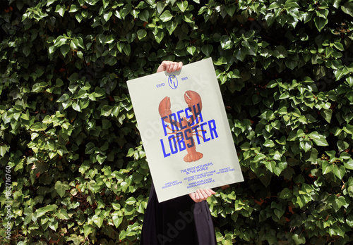 Mockup of person holding customizable vertical A2 poster by hedge