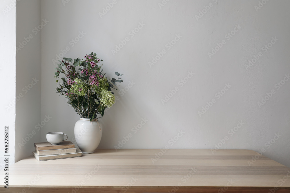 Elegant Scandinavian interior. Vase with floral bouquet. Eucalyptus tree branches, hydrangea, wax flowers. Cup of tea, cofffe. Old books. Empty wall background. Modern living room, home office.