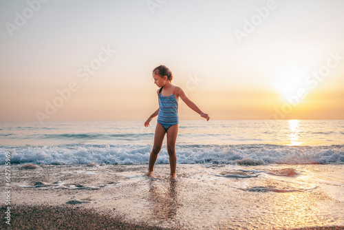 The child happily plays in the waves on the seashore. A girl in a blue swimsuit laughs while playing in the waves on the beach © BigBlueStudio