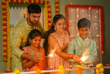Happy parents with kids playing with diwali sparklers or Crackers by holding hands at home during festive celebration - concept of childhood memories, family bonding and traditional culture