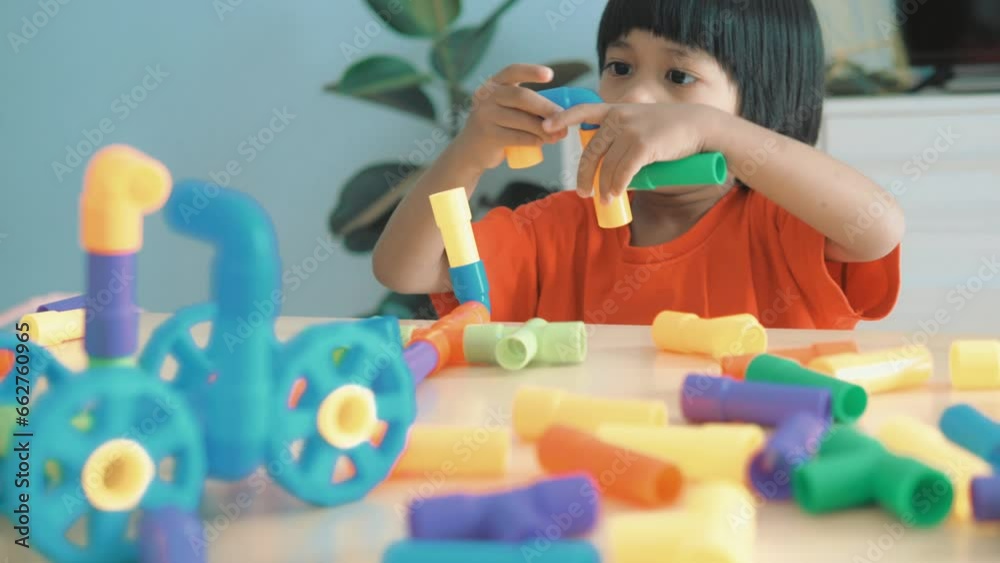Adorable Asian boy, about 3-6 years old, wearing a pink shirt, happily playing with a puzzle shaped like a water pipe and having various toy items placed on a table inside his home, white background. Adorable Asian boy, about 3-6 years old, wearing a pink shirt, happily playing with a puzzle shaped like a water pipe and having various toy items placed on a table inside his home, white background.