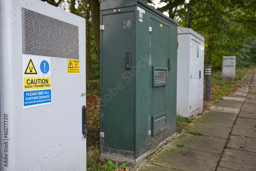 A row of telecommunications cabinets are lined up beside a pavement.  One of the cabinets in the foreground has stickers warning about radio transmissions and high electricity voltages