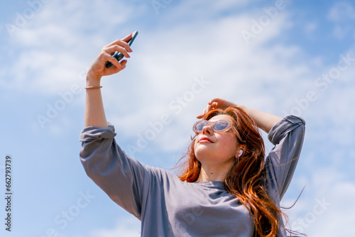 Tableau sur toile a young red-haired girl in sunglasses with a phone in her hands takes a photo of