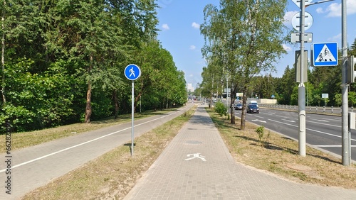 Tile-paved bike and pedestrian paths, marked with signs and pavement markings, are located between the park's trees and the roadway of the city street where cars travel. Sunny summer weather