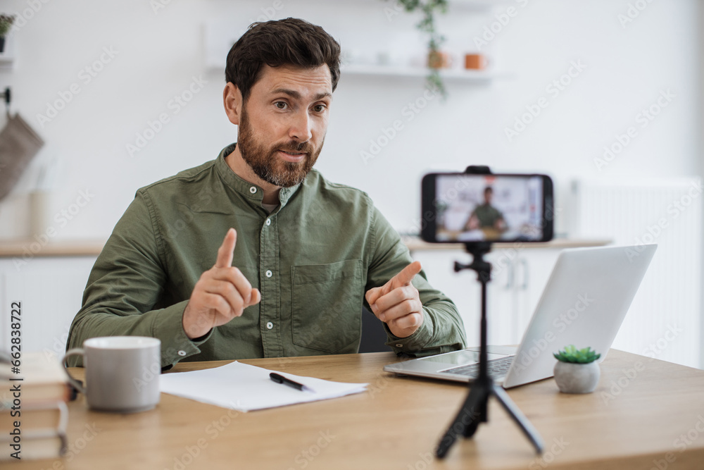 Modern smartphone fixed on tripod filming handsome caucasian man in casual wear working on portable laptop at home office. Blurred background. Technology, people and blogging concept. Modern smartphone fixed on tripod filming handsome caucasian man in casual wear working on portable laptop at home office. Blurred background. Technology, people and blogging concept.