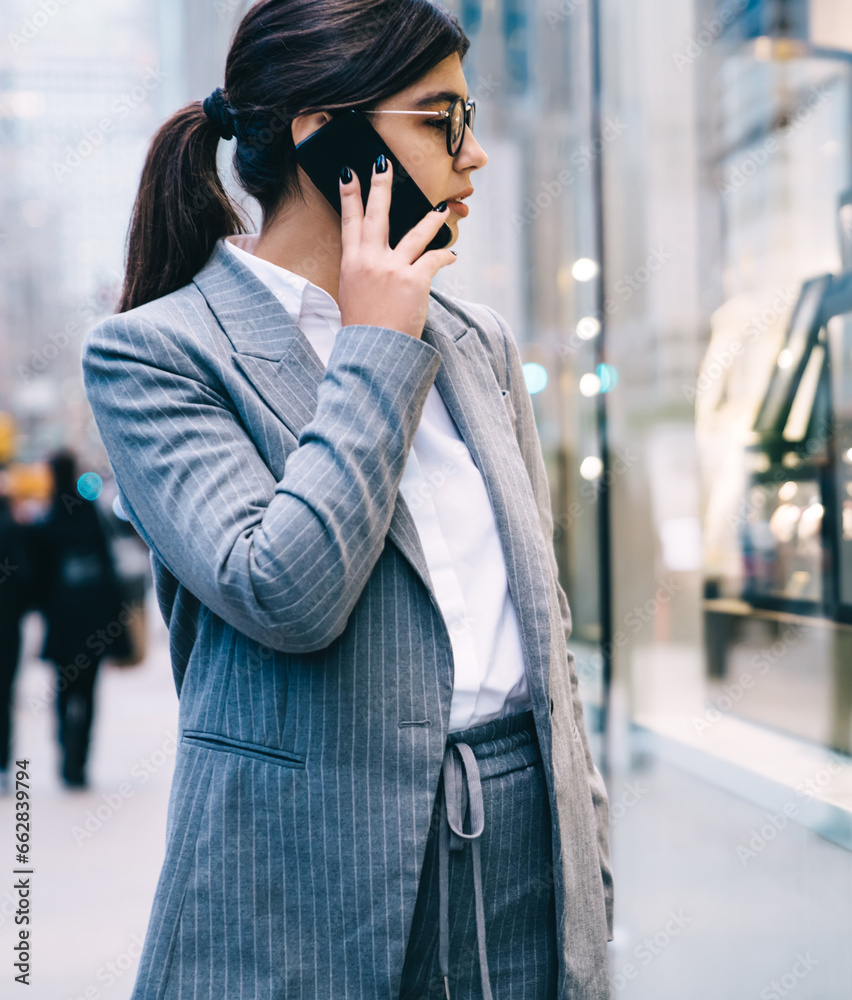 Fototapeta premium Confident formal young beautiful business lady talking on smartphone while walking on street