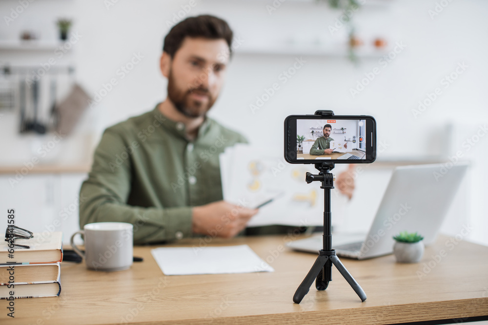 Blur background of caucasian man sitting at desk with laptop and showing financial report on smartphone camera. Competent financier making video lesson about reading and interpreting graphic charts. Blur background of caucasian man sitting at desk with laptop and showing financial report on smartphone camera. Competent financier making video lesson about reading and interpreting graphic charts.