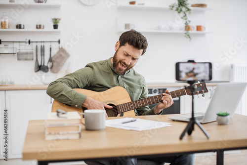 Inspired caucasian man virtuoso playing guitar while sitting at desk and recording video on modern smartphone with tripod. Male vlogger creating new lesson about instrument techniques to social media.