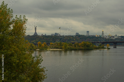 Autumn landscape of Dnipro River in Kyiv during cloudy day. Famous Kyiv's hills on right bank of river.Kyiv Pechersk Lavra, residential buildings and Motherland Monument on horizon.Tree leaves border