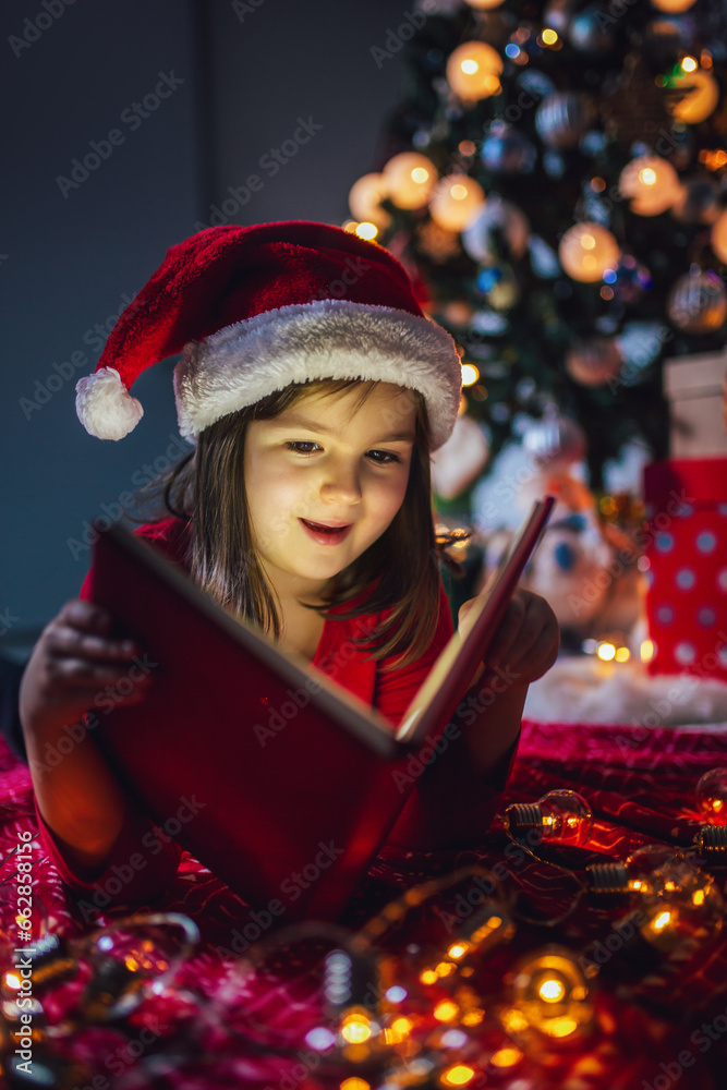 Little girl is reading a book on Christmas. Colorful lights, Santa Claus hat and christmas tree