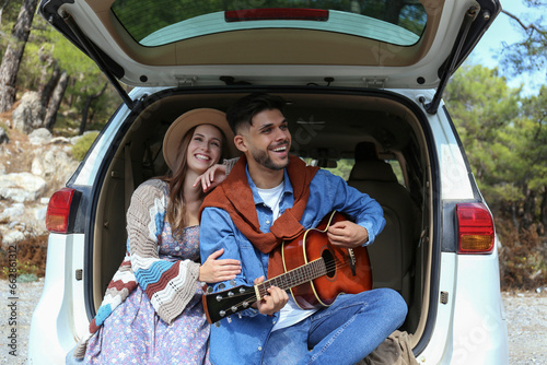 Happy hipster couple sitting in the trunk of their car and singing a songs to the acoustic guitar. Close up, copy space, background. © Evrymmnt