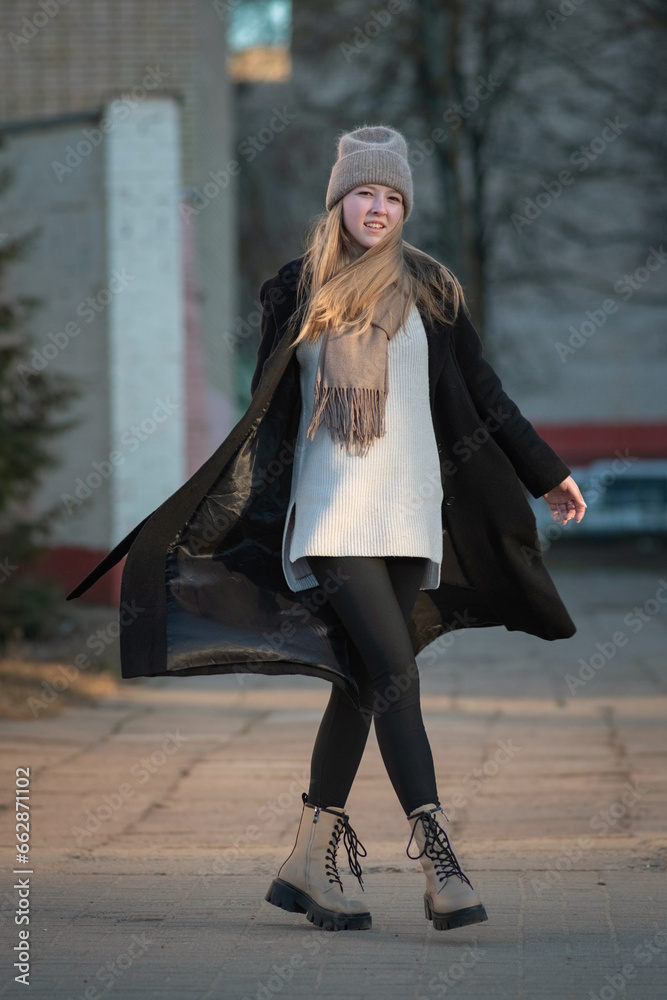 Portrait of a young beautiful blonde girl in a knitted hat and dark coat in an urban environment.