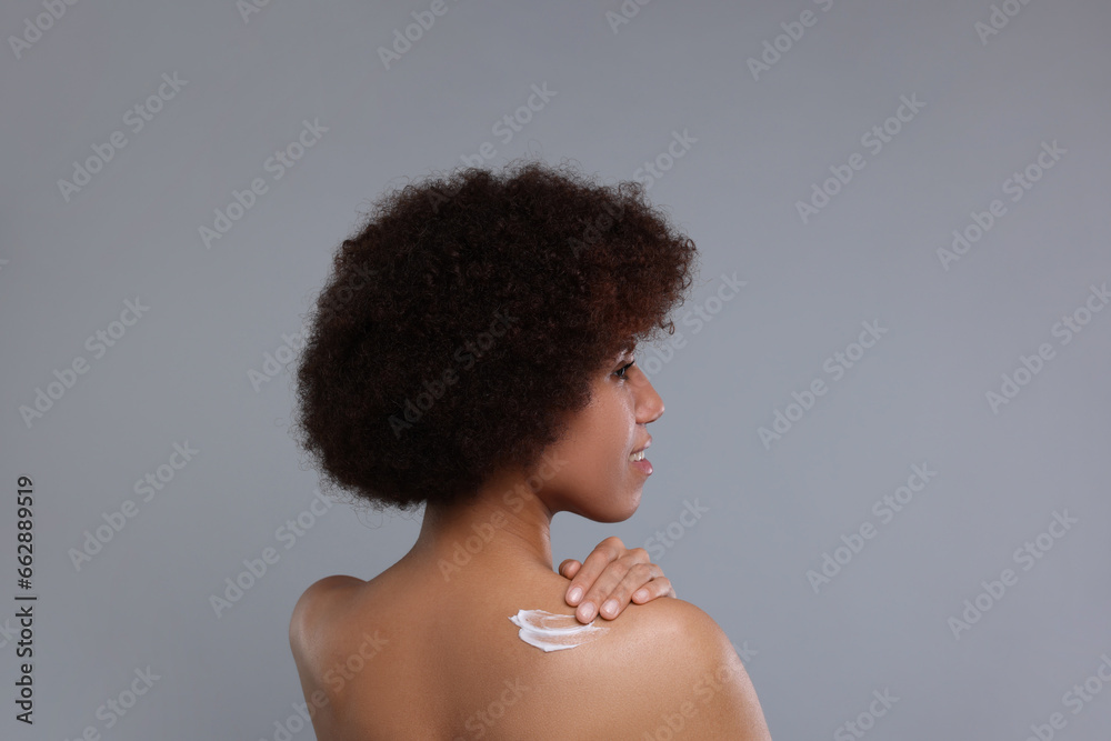 Beautiful young woman applying body cream onto back on grey background Beautiful young woman applying body cream onto back on grey background