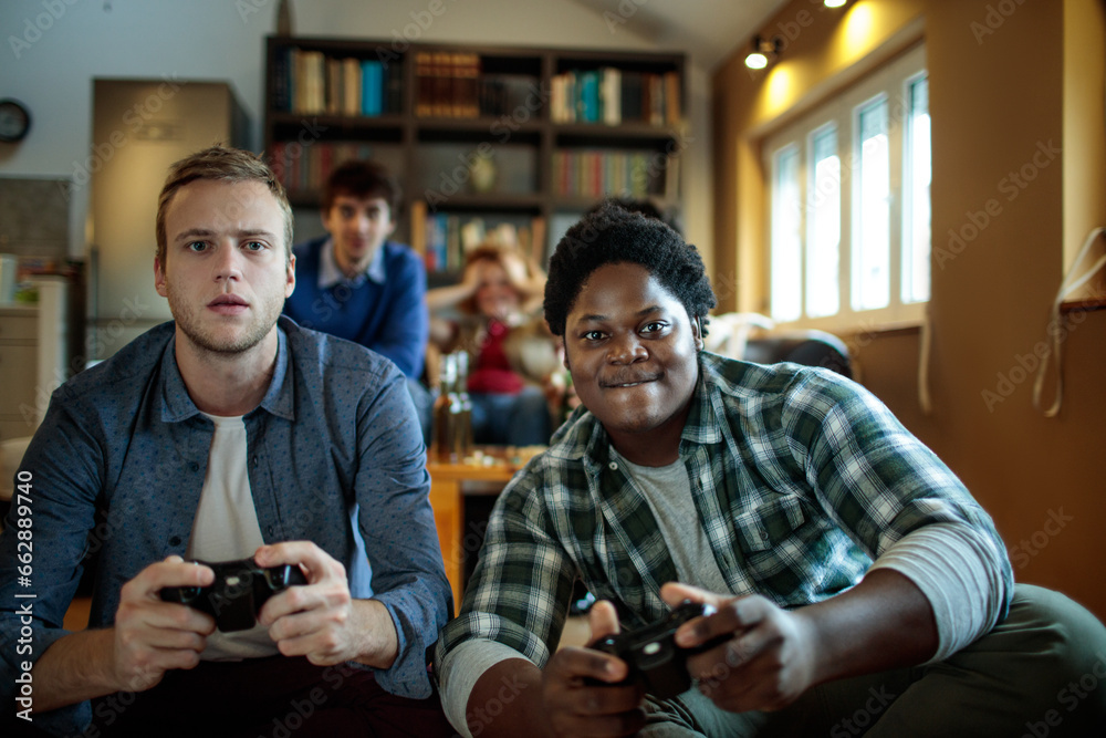 Two diverse male friends playing video games with a controller in the living room at home Two diverse male friends playing video games with a controller in the living room at home