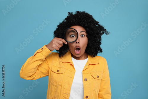 Emotional woman looking through magnifier glass on light blue background