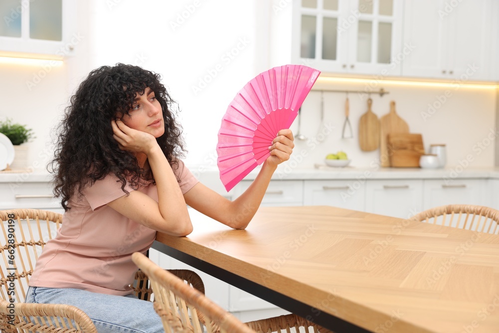 Obraz premium Young woman waving pink hand fan to cool herself at table in kitchen. Space for text