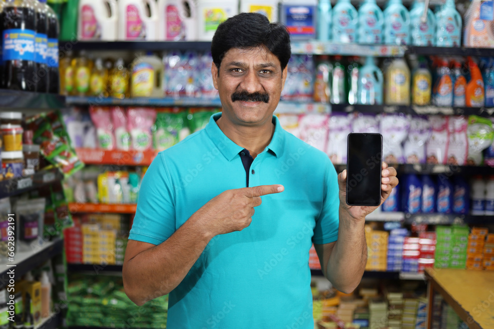 Fototapeta premium Smiling mid aged man purchasing in a grocery store pointing towards the mobile app. Buying grocery in supermarket pointing towards the mobile app.