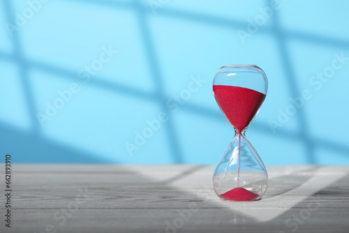 Hourglass with red flowing sand on white wooden table against light blue background, space for text