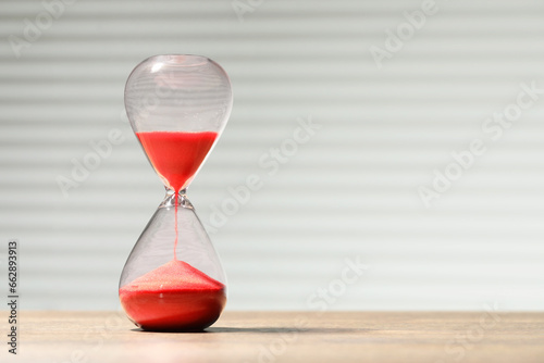 Hourglass with red flowing sand on wooden table against light background, space for text