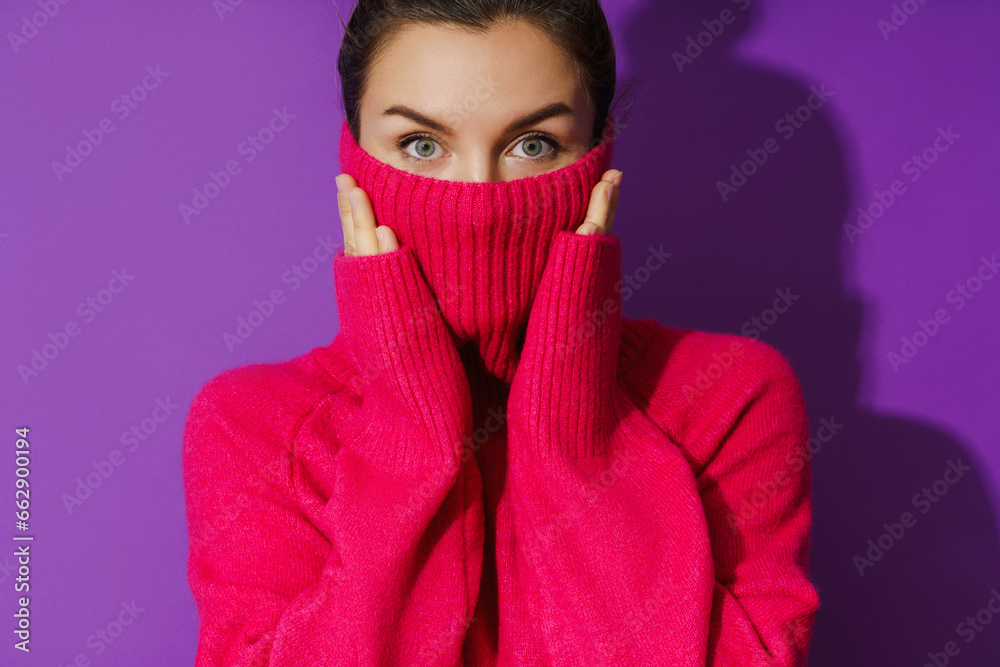 Young woman is hiding her face inside a warm and cozy polo neck sweater against purple background Young woman is hiding her face inside a warm and cozy polo neck sweater against purple background