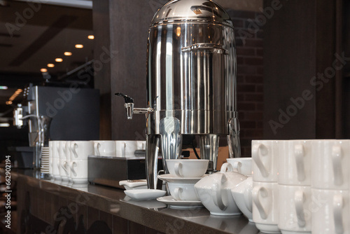 Large electric kettle boiler with hot water for tea, cups and mugs on the bar prepared for self-service breakfast buffet.