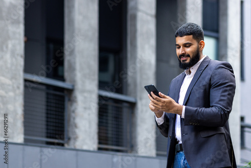 Young muslim man businessman standing on the street near an office building and using the phone, going to a meeting, calling, typing a message