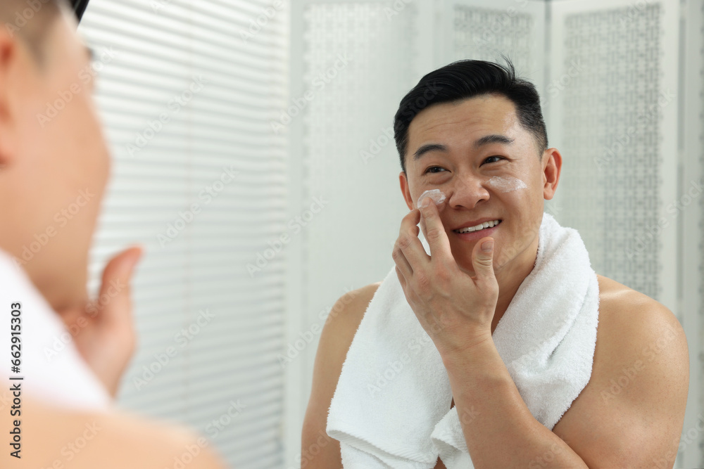 Handsome man applying cream onto his face near mirror indoors Handsome man applying cream onto his face near mirror indoors