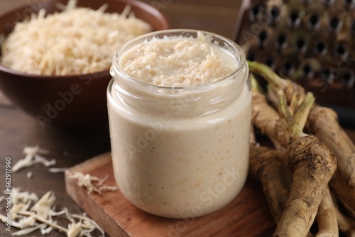 Spicy horseradish sauce in jar and roots on table, closeup