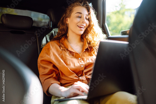 A young curly woman with a laptop sitting in the back seat of a car. Beautiful woman uses laptop in car. Remote work, freelance. © maxbelchenko