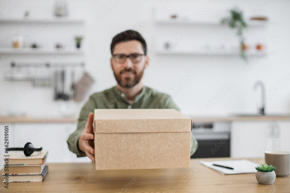 View from smartphone camera of smiling bearded man in eyeglasses showing cardboard box with online order while sitting at desk. Popular vlogger filming new video of technology unpacking at home. View from smartphone camera of smiling bearded man in eyeglasses showing cardboard box with online order while sitting at desk. Popular vlogger filming new video of technology unpacking at home.