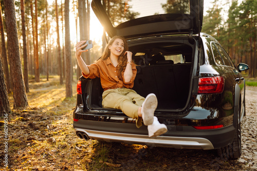 Stylish woman takes a selfie, communicates via video call from a smartphone in the trunk of a car at sunset. Autumn trip. A young woman uses the phone and travels by car. © maxbelchenko