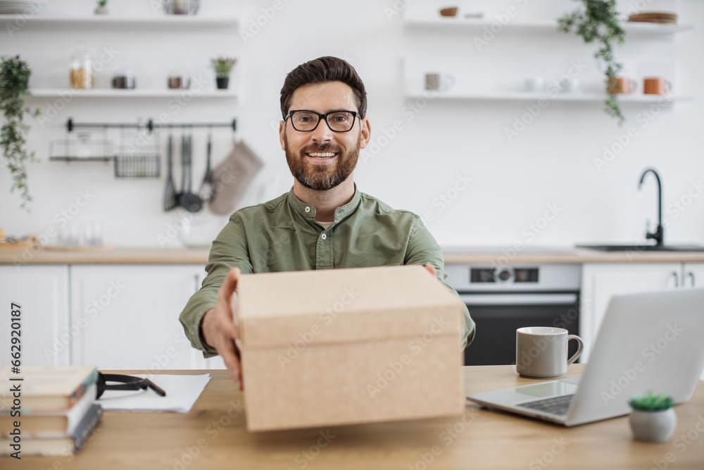 View from smartphone camera of smiling bearded man in eyeglasses showing cardboard box with online order while sitting at desk. Popular vlogger filming new video of technology unpacking at home. View from smartphone camera of smiling bearded man in eyeglasses showing cardboard box with online order while sitting at desk. Popular vlogger filming new video of technology unpacking at home.