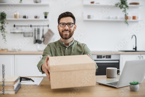 View from smartphone camera of smiling bearded man in eyeglasses showing cardboard box with online order while sitting at desk. Popular vlogger filming new video of technology unpacking at home.