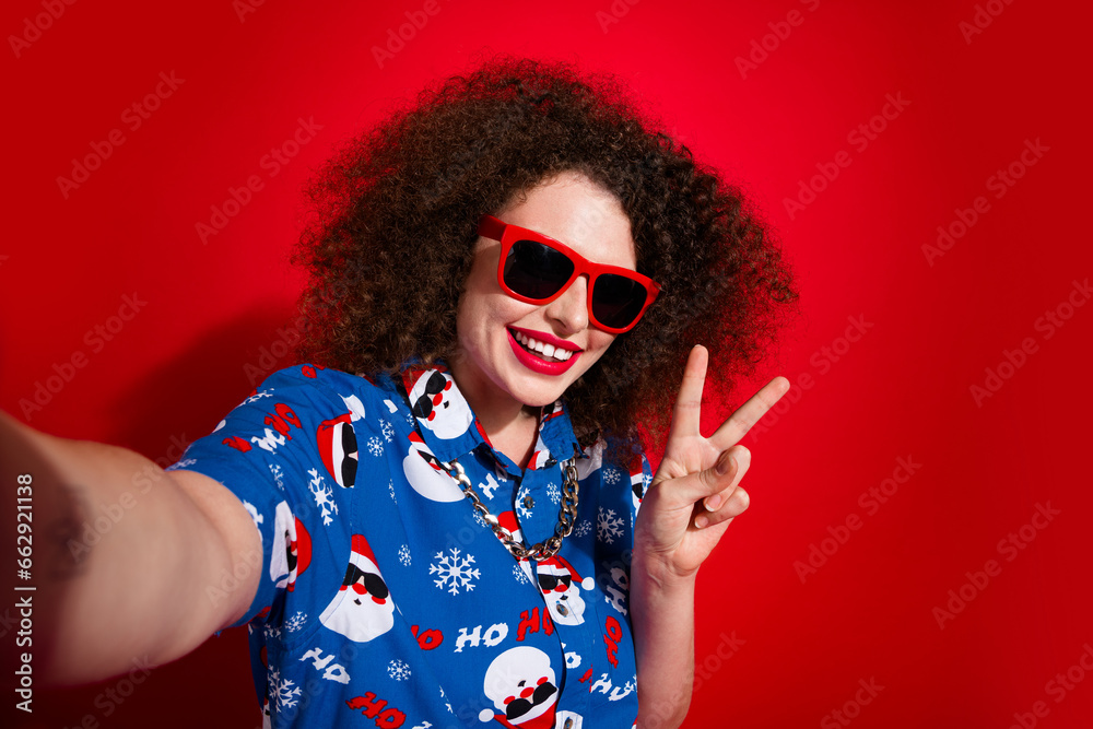 Photo of positive pretty woman wear ugly santa claus print x-mas shirt dark spectacles recording video v-sign isolated red color background