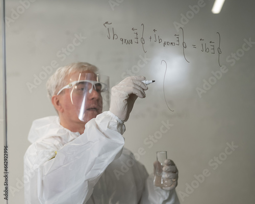 An elderly Caucasian male chemist in a protective suit writes on glass. 