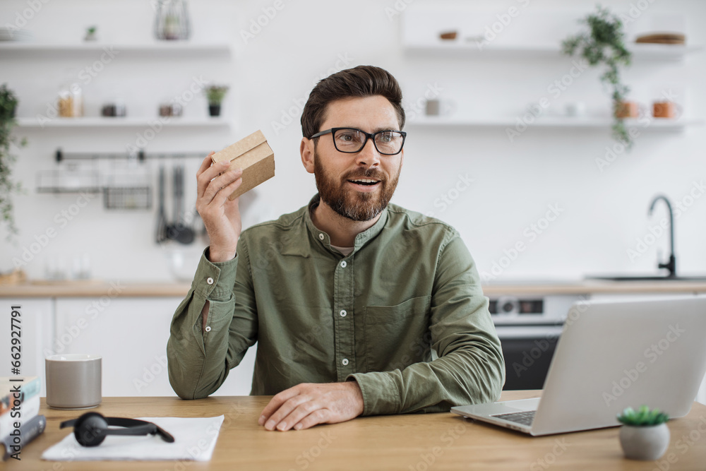 Caucasian bearded man in eyeglasses and looking at small cardboard parcel with focused facial expression. Social media influencer streaming unpacking process of online order from home. - obrazy, fototapety, plakaty 