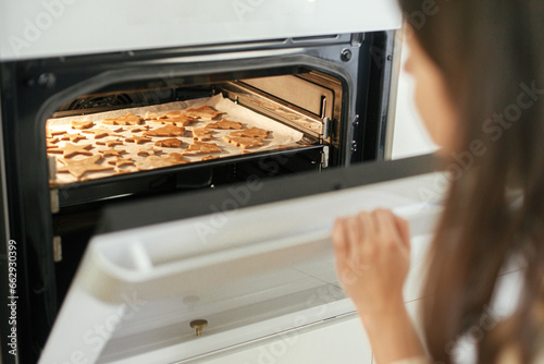 Making christmas gingerbread cookies. Woman putting tray with fresh gingerbread cookies in christmas festive shapes in modern oven in white kitchen