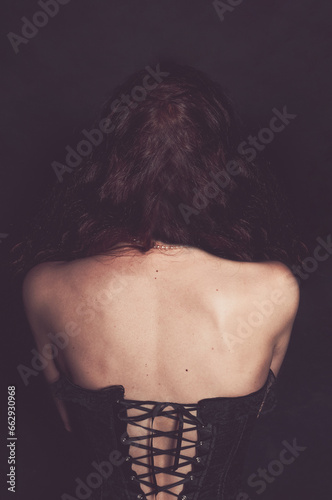 The bare back of a girl dressed in a black corset, a pearl necklace around her neck, and dark curly hair. Gothic clothing style.