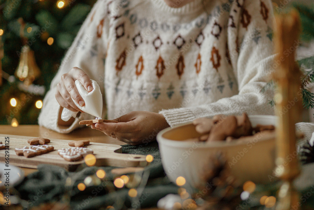 Hands decorating christmas gingerbread cookies with icing on rustic wooden table on background of golden lights. Atmospheric Christmas holiday traditions. Decorating cookies with sugar frosting