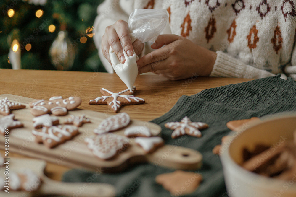 Hands decorating christmas gingerbread cookies with icing on rustic wooden table close up. Atmospheric Christmas holiday traditions. Decorating baked cookies with sugar frosting. Family time - obrazy, fototapety, plakaty 