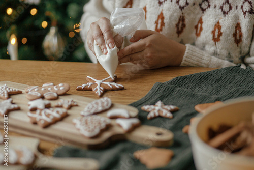 Hands decorating christmas gingerbread cookies with icing on rustic wooden table close up. Atmospheric Christmas holiday traditions. Decorating baked cookies with sugar frosting. Family time