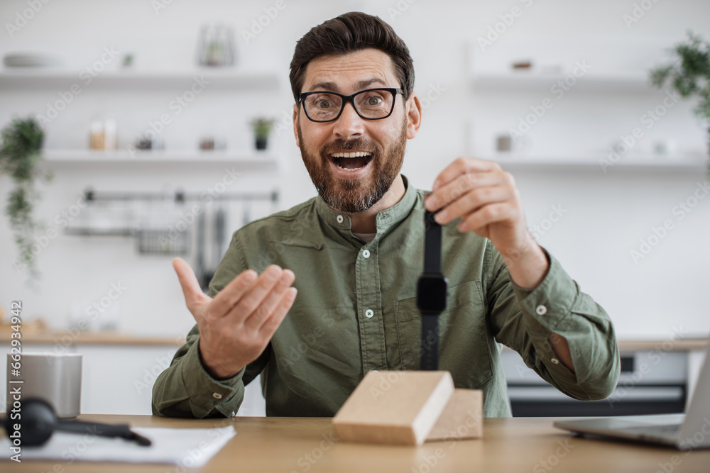 Caucasian male influencer wearing eyeglasses recording video blog about unboxing of new smart watch. Bearded young guy doing review of modern gadget while working from home. Caucasian male influencer wearing eyeglasses recording video blog about unboxing of new smart watch. Bearded young guy doing review of modern gadget while working from home.