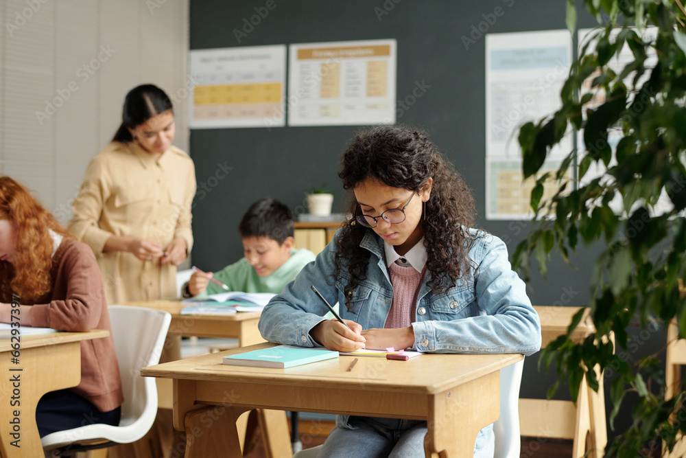 Diligent multiethnic schoolgirl making notes in copybook during individual work at lesson while teacher checking assignment of schoolboy Diligent multiethnic schoolgirl making notes in copybook during individual work at lesson while teacher checking assignment of schoolboy