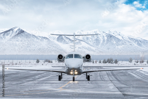 Front view of the white business jet taxiing on airport taxiway in winter on the background of high scenic snow capped mountains