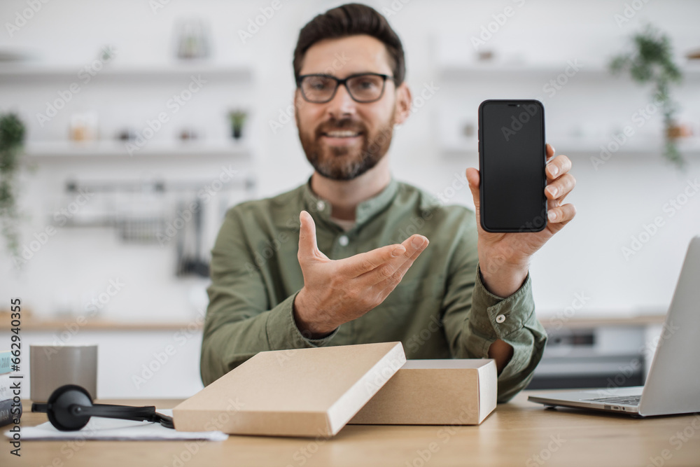 First person view of male influencer sharing feedback with followers while live streaming unpacking of modern smartphone. Bearded man sitting at desk and pointing with hand on new cell phone. First person view of male influencer sharing feedback with followers while live streaming unpacking of modern smartphone. Bearded man sitting at desk and pointing with hand on new cell phone.