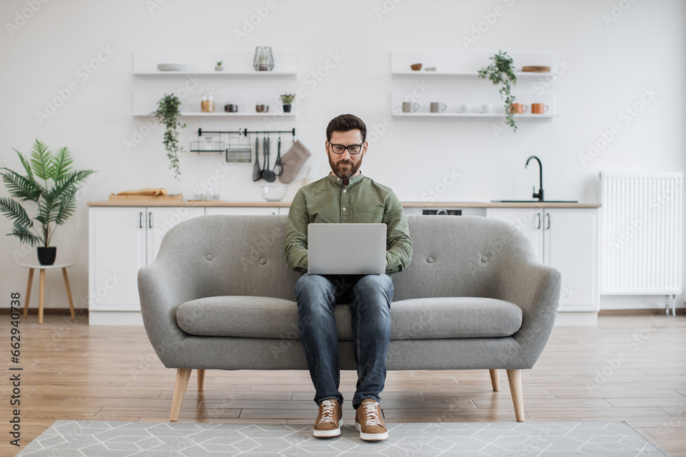 Obraz premium Focused caucasian man dressed in casual attire typing on wireless laptop while sitting on comfy sofa. Attractive male freelancer in eyeglasses doing online work while staying at home.