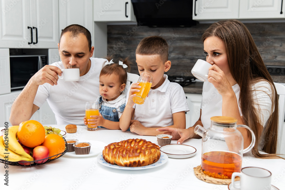 Portrait Of Happy European family Of Four Having Breakfast at home In Kitchen Together. Parents With children Enjoying Tasty Food And Drinking Orange Juice While Sitting At Table