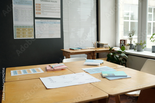 Part of spacious classroom with square wooden desk standing in the center against window, wall with grammar tables, green plants and other stuff